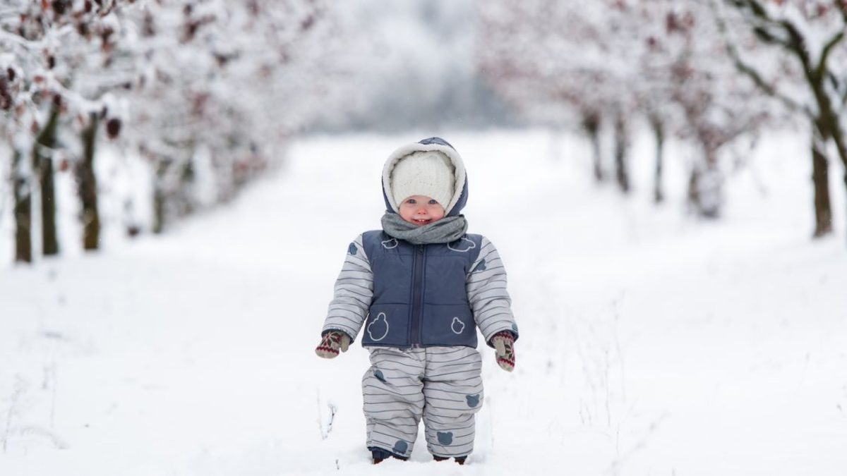 A bundled-up toddler in a winter snowsuit and hat walking through a snowy path