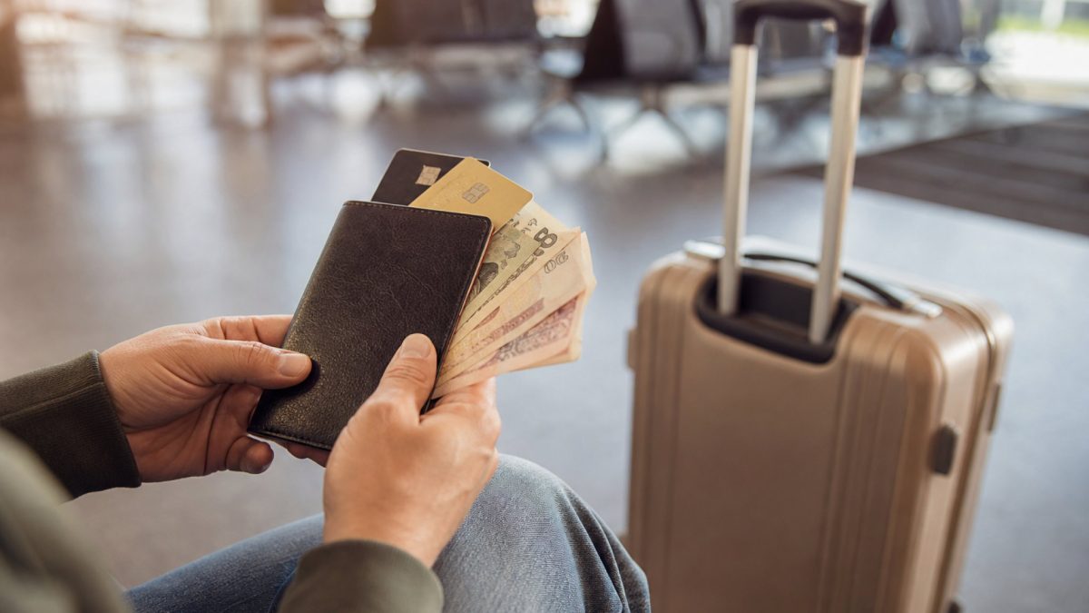 Traveler holding a wallet with credit cards and foreign cash beside a suitcase at an airport gate