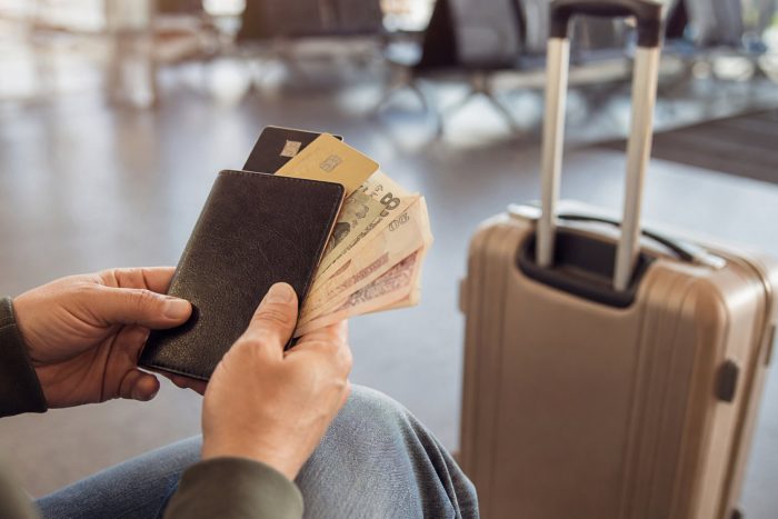 Traveler holding a wallet with credit cards and foreign cash beside a suitcase at an airport gate